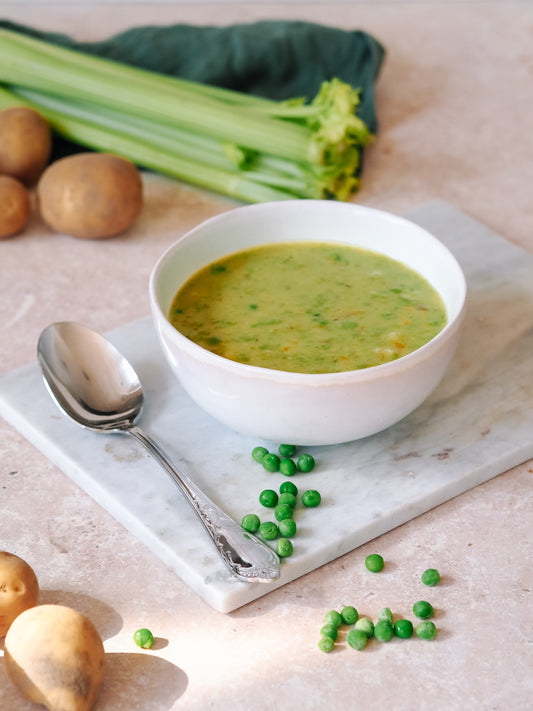 Cremige Erbsensuppe in einer weißen Schüssel auf einem Marmoruntersetzer, begleitet von einem silbernen Löffel und verstreuten grünen Erbsen, mit frischen Zutaten wie Sellerie und Kartoffeln im Hintergrund