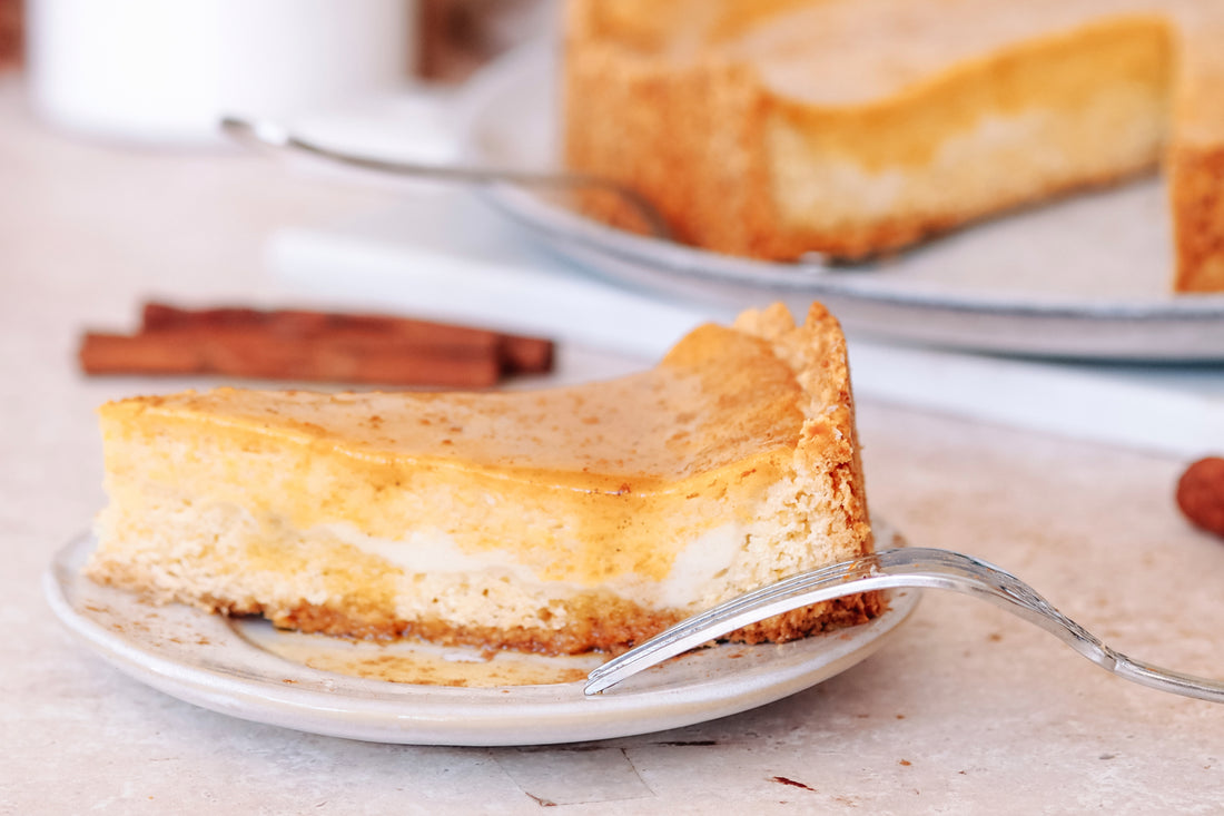 Ein Stück veganer Cheesecake auf einem weißen Teller, mit einer Gabel daneben und einem weiteren Cheesecake im unscharfen Hintergrund, neben einem Zuckerbehälter und Zimtstangen auf einer hellen Marmoroberfläche