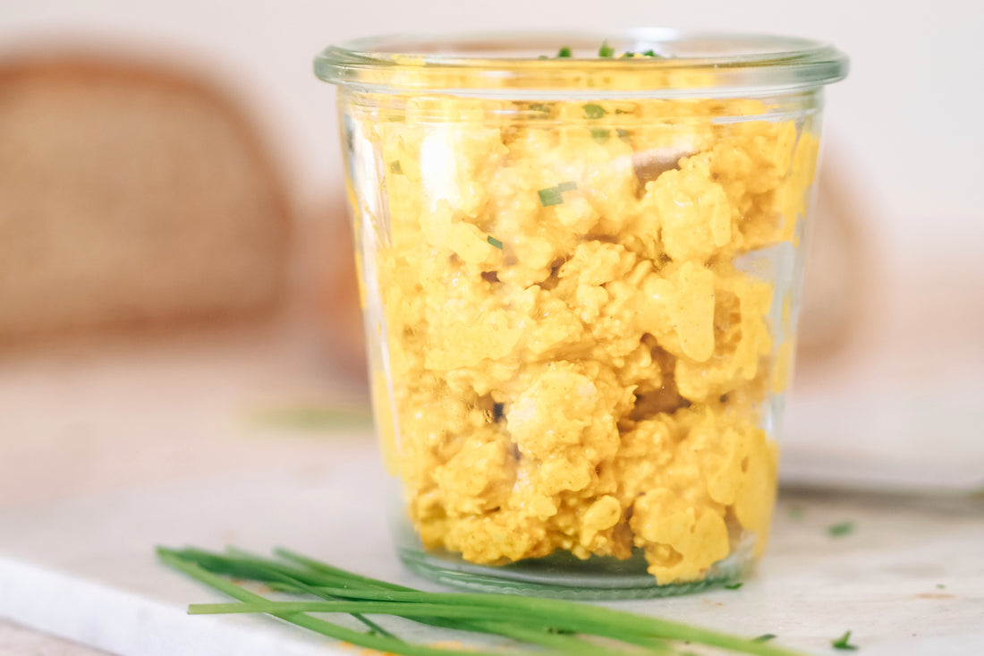 Veganer Eiersalat in einem durchsichtigen Glasgefäß, garniert mit frischen Schnittlauchröllchen, auf einem Marmoruntergrund mit einer Spur von Gewürzen und einem unscharfen Brotlaib im Hintergrund