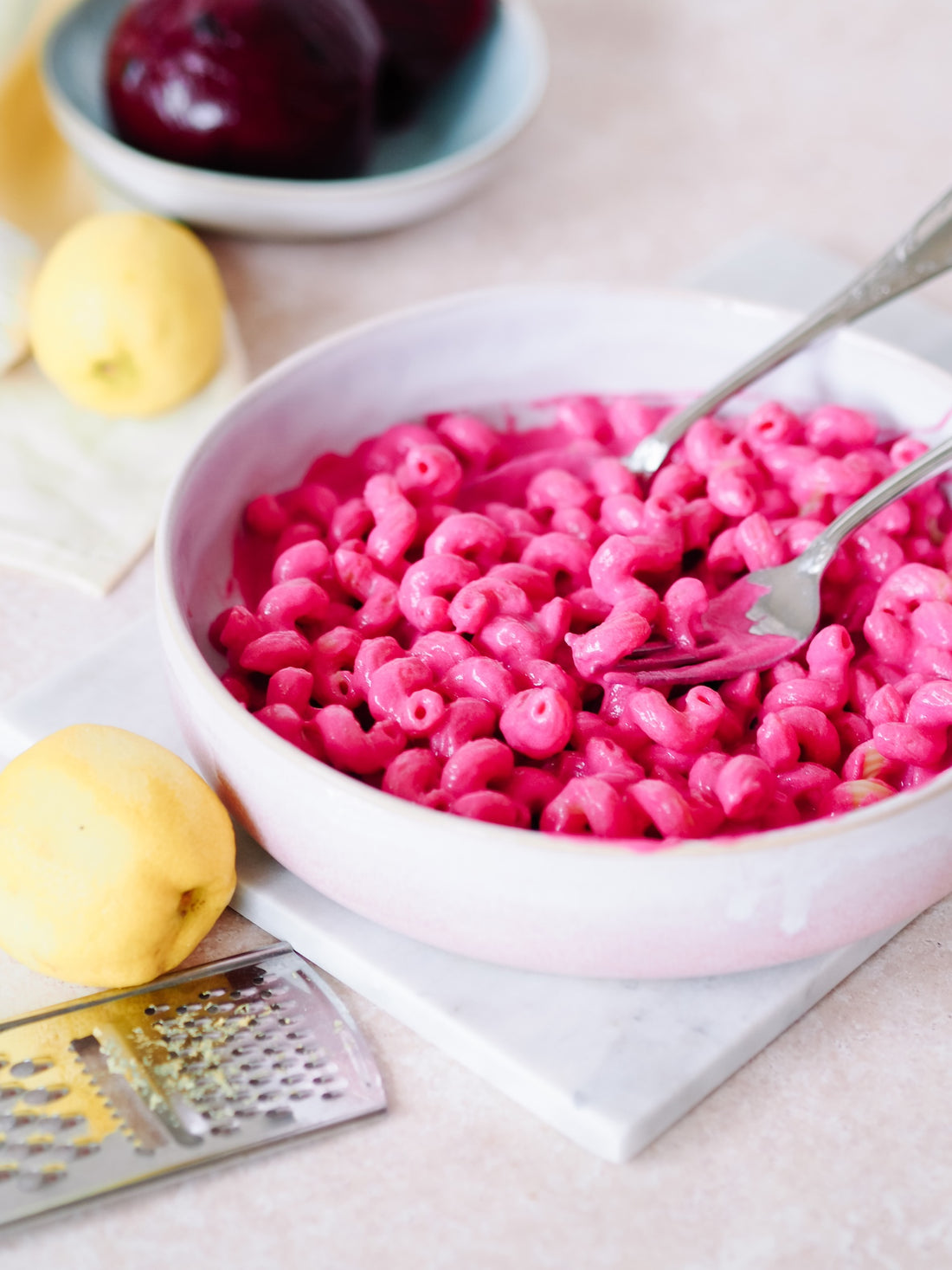 Bunte, gekochte Rote Beete Nudeln in einer weißen Schüssel mit Zitronen und einer Reibe auf Marmoruntergrund